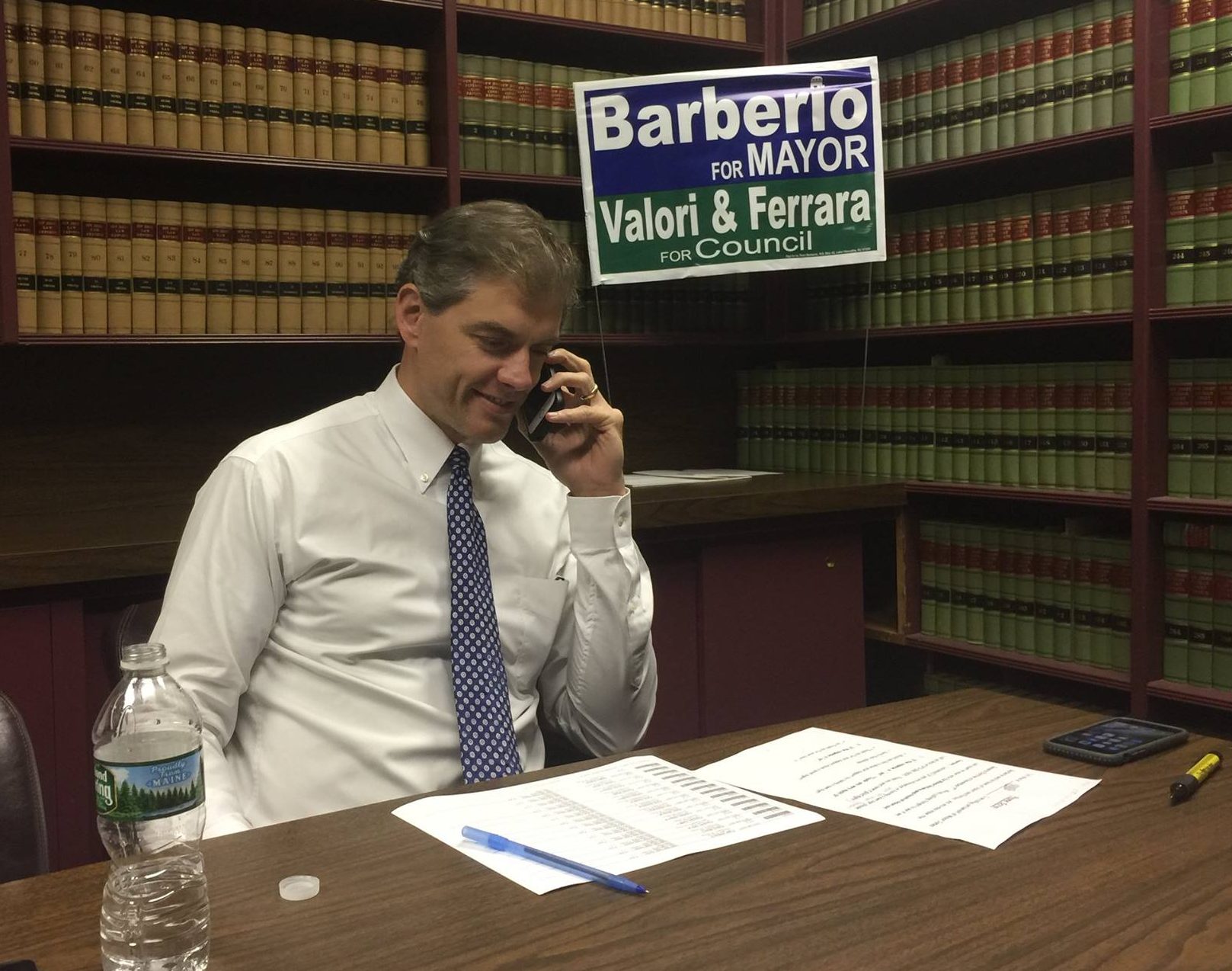 Assemblyman Jay Webber, phone in hand and smiling mid-sentence, in a Parsippany law office with a Barberio for Mayor / Valori & Ferrara for Council sign on the law-library shelves behind him and two sheets on the desk in front of him, a call-sheet grid and a script.